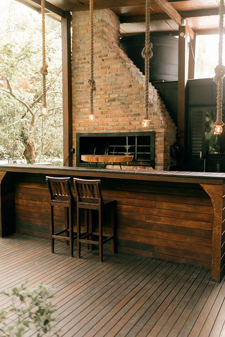 Cucina outdoor con caminetto in mattoni a vista, bancone in legno scuro, sgabelli in legno e luci a incandescenza su fune, in ambiente coperto con pareti vetrate e vegetazione
