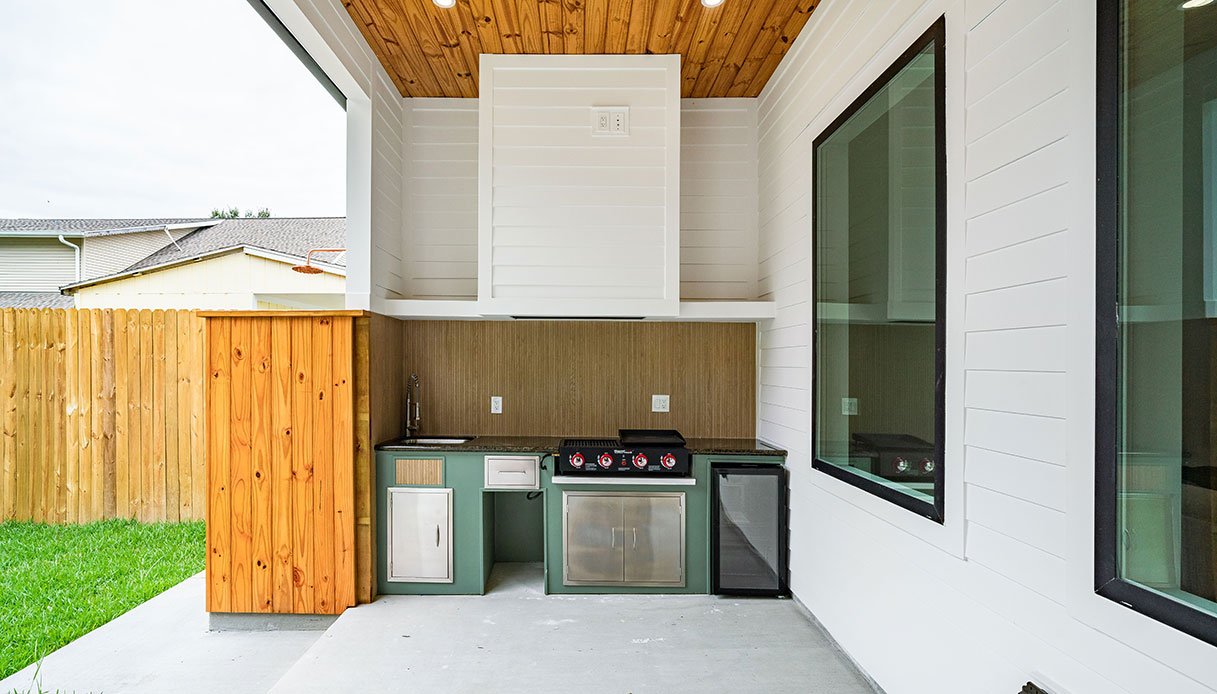 Cucina outdoor modulare con ante in verde salvia, cappa in acciaio bianco, griglia a gas e frigorifero da incasso su patio coperto con soffitto in legno