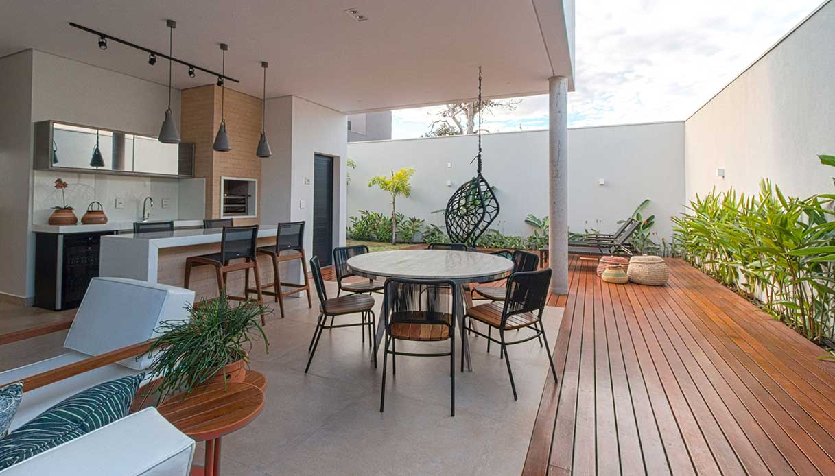 Cucina outdoor con bancone in acciaio e top scuro, zona pranzo con tavolo rotondo e sedie in metallo nero, su terrazza con deck in legno e giardino con piante tropicali