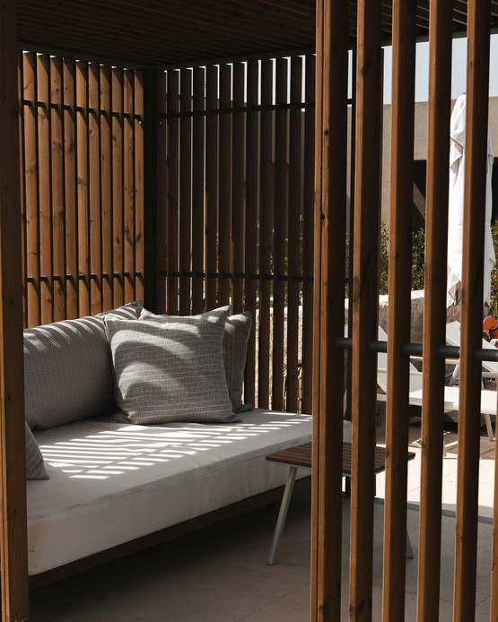 Daybed in legno con cuscini grigi all'interno di una cabana a doghe verticali, vista su una terrazza con ombrellone