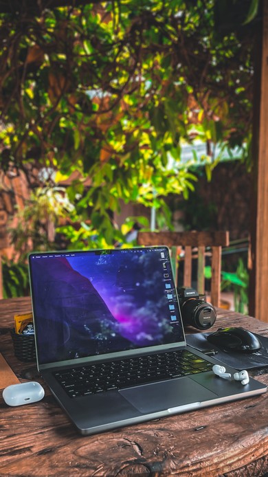 Laptop aperto su un tavolo in legno immerso nel verde, con fotocamera, cuffie wireless e mouse
