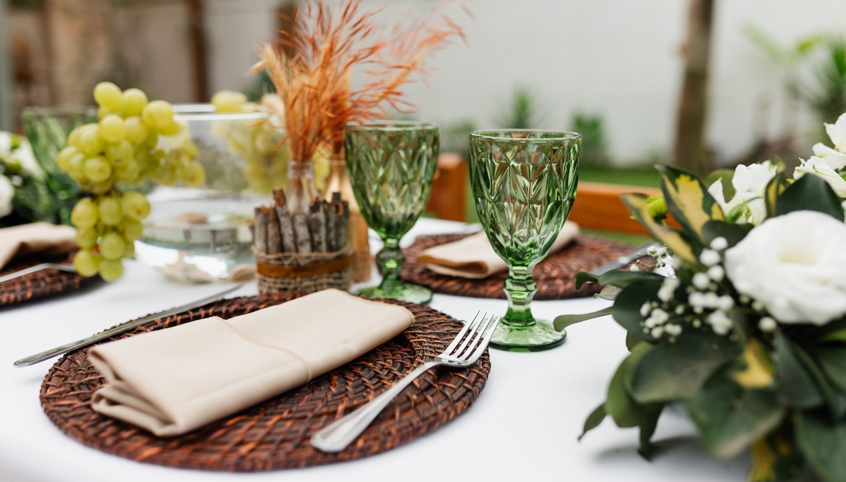 Primo piano di una mise en place con sottopiatto in rattan, tovagliolo beige, calici in vetro lavorato verde e grappoli d'uva