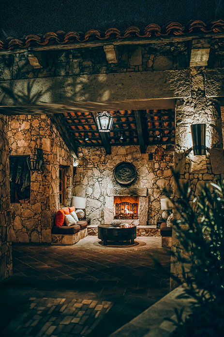 Patio rustico in pietra con camino acceso, lanterna a soffitto e divanetto con cuscini in un'atmosfera notturna