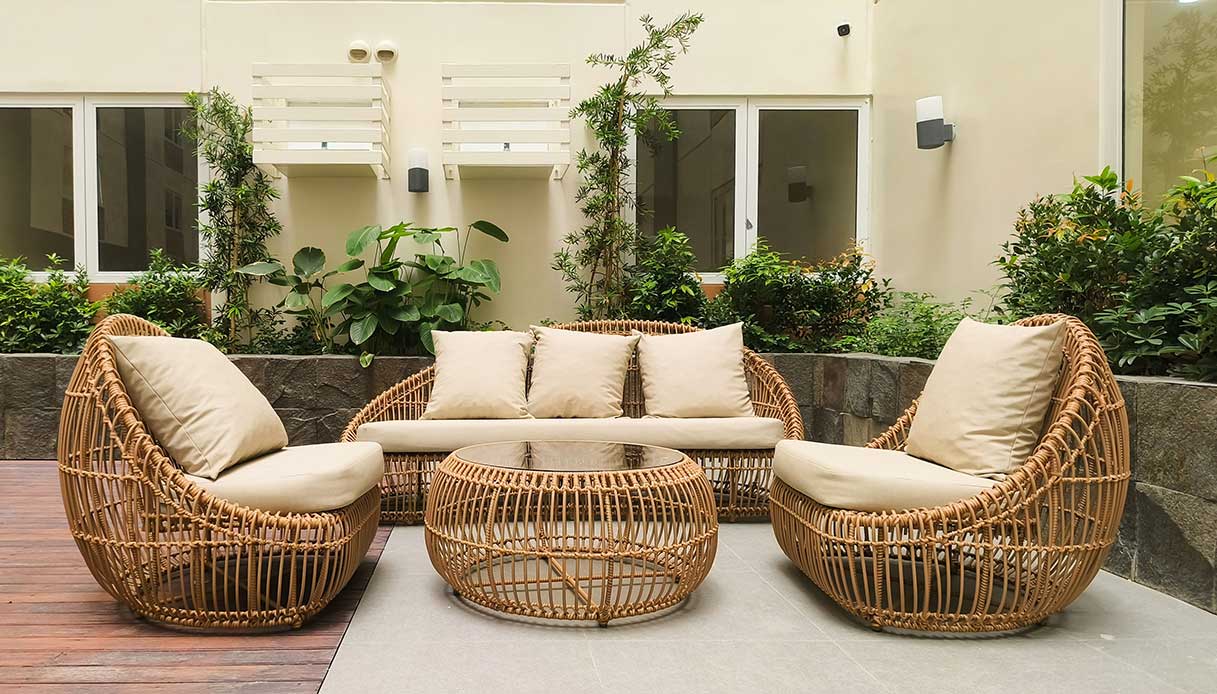 Salotto outdoor con poltrone e divano in rattan intrecciato, cuscini beige e tavolino basso con piano in vetro