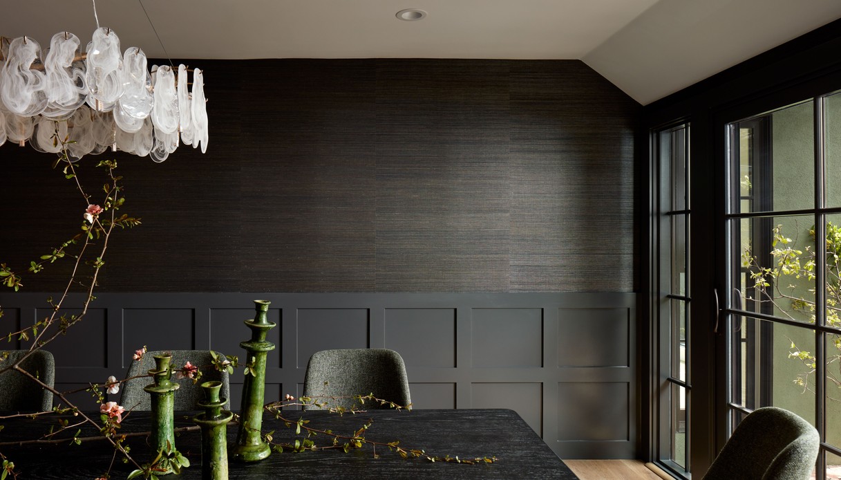Sala da pranzo con pareti scure in carta da parati grasscloth, tavolo nero con candelieri verdi in ceramica, sedie verde salvia e lampadario in vetro soffiato