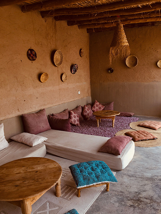 Salotto in stile marocchino con pareti in terracotta, cesti e piatti intrecciati appesi, cuscini bordeaux e pouf blu