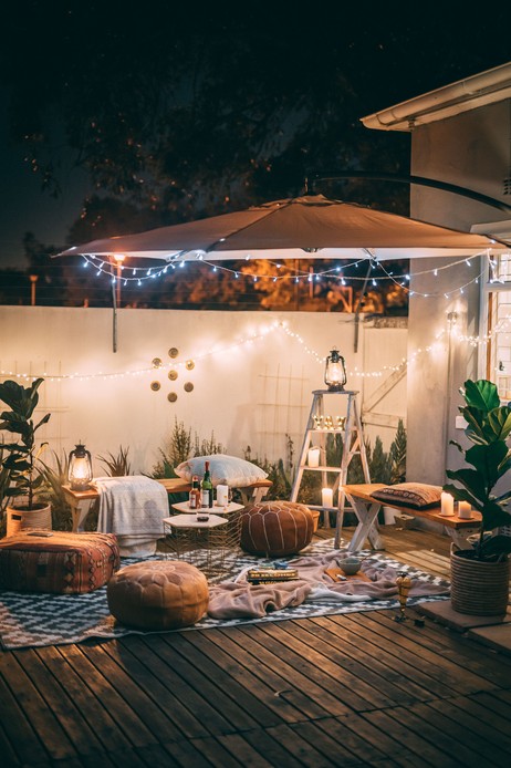 Terrazza allestita con pouf in pelle, cuscini, plaid su tappeto a motivi geometrici, lanterne e ombrellone con lucine