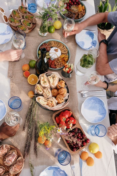Tavola estiva vista dall'alto con ceramiche dipinte in blu, bicchieri azzurri, runner in juta e vassoi di frutta e pane, mani che reggono i calici
