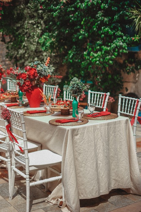 Tavola apparecchiata all'aperto con tovaglia in lino stropicciato, sottopiatti in rattan intrecciato e tovaglioli rossi annodati