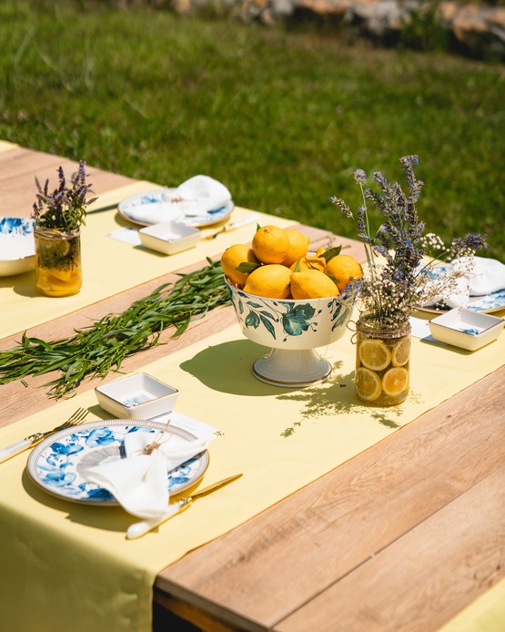 Tavolo in legno con runner giallo, alzata di limoni, ceramiche bianche e blu a motivo floreale e mazzetti di lavanda in barattoli di vetro