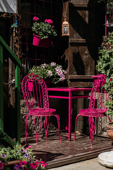 Tavolino e due sedie in ferro battuto rosa fucsia su una veranda, con gerani rosa e lanterna appesa