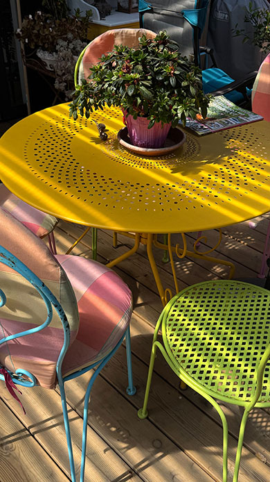 Tavolo giallo perforato con sedie in metallo di colori diversi, cuscini a quadretti e righe, pianta in vaso al centro