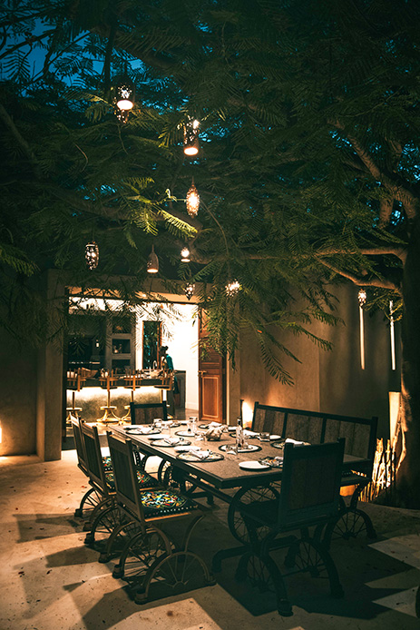 Tavolo da pranzo apparecchiato in un patio notturno sotto un grande albero decorato con lanterne sospese e luci d'accento sulle pareti