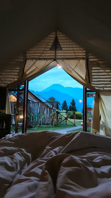 Vista dall'interno di una tenda glamping verso un giardino recintato e montagne blu al crepuscolo, letto in primo piano