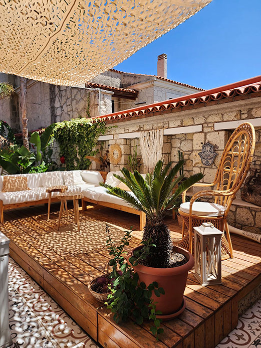 Terrazza in pietra con tenda ombreggiante traforata, divano bianco, poltrona in rattan a forma di pavone e palma in vaso di terracotta