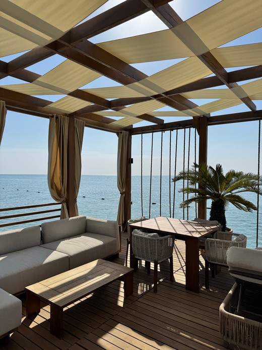 Terrazza coperta da vele ombreggianti in tessuto, con divano, tavolo da pranzo e palma, affacciata sul mare