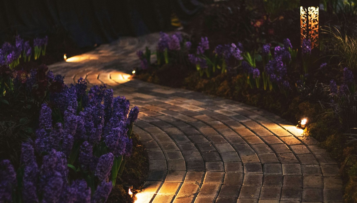 Vialetto curvo in pietra illuminato di sera da faretti a incasso, con aiuole di giacinti viola ai lati