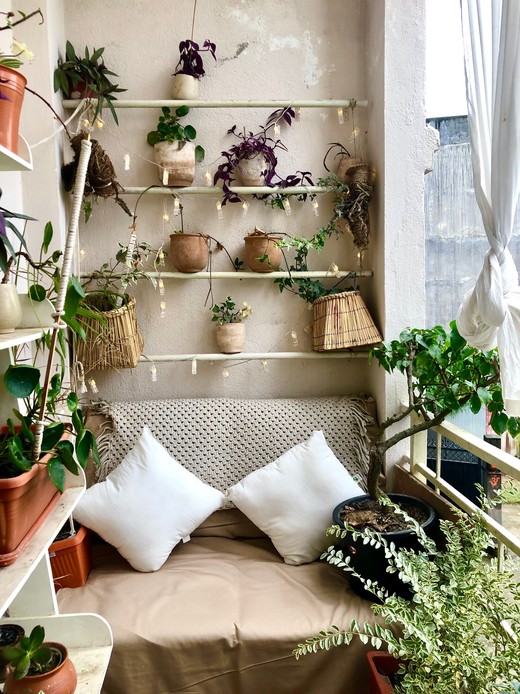 Angolo di balcone con materasso da terra, cuscini bianchi e plaid intrecciato, sotto mensole cariche di piante in vasi di terracotta e lucine