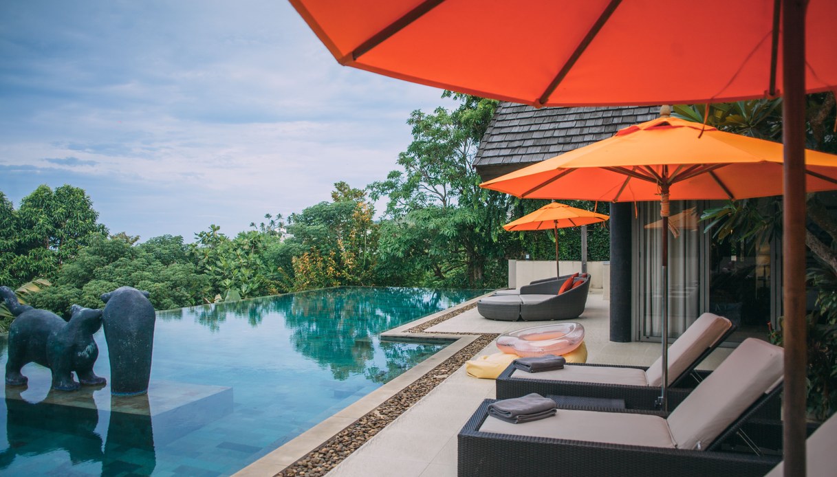 Bordo di una infinity pool con lettini in rattan scuro, ombrelloni arancioni e vegetazione tropicale sullo sfondo