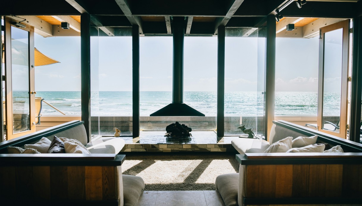 Interno di una casa sul mare con ampie vetrate, panche in legno con cuscini chiari, camino sospeso nero e vista diretta sull'oceano.