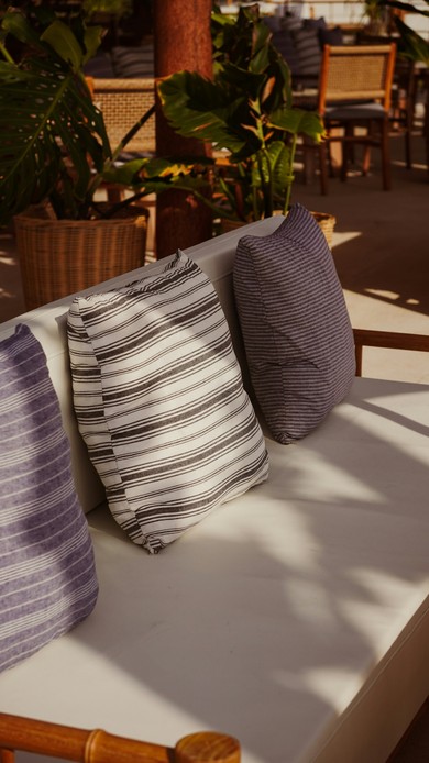 Cuscini a righe in tessuto effetto lino appoggiati a un divano bianco su una terrazza, con piante tropicali sullo sfondo