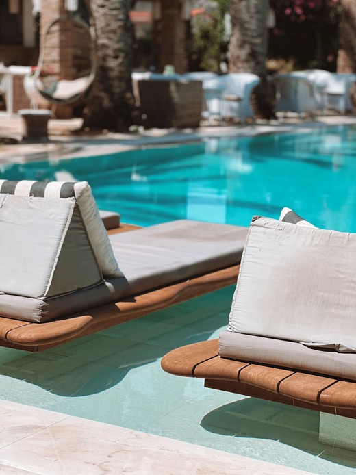 Lettini prendisole con struttura in legno di teak e cuscini chiari, posizionati a pelo d'acqua lungo il bordo di una piscina