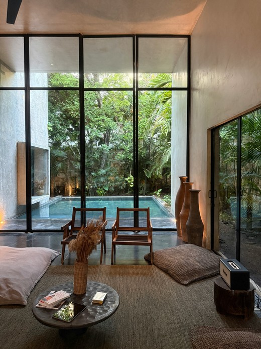 Salotto interno con cuscini da terra, sedute in canna e una parete vetrata a tutta altezza affacciata su piscina e giardino tropicale