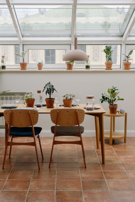 Sala da pranzo sotto una veranda vetrata con pavimento in cotto, sedie in legno, vasi in terracotta e piante da interno