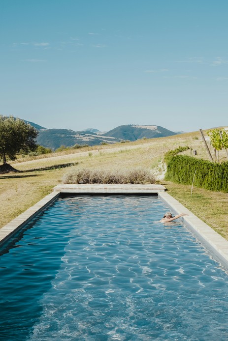 Lunga piscina a sfioro in un paesaggio collinare con un olivo, una persona che nuota e i monti sullo sfondo