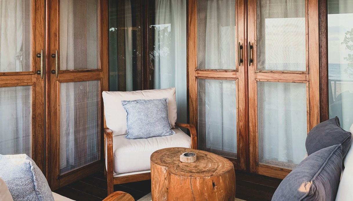 Angolo salotto con poltrone in teak, cuscini bianchi e blu, tavolino ricavato da un tronco e ampie finestre in legno con vista sul mare.