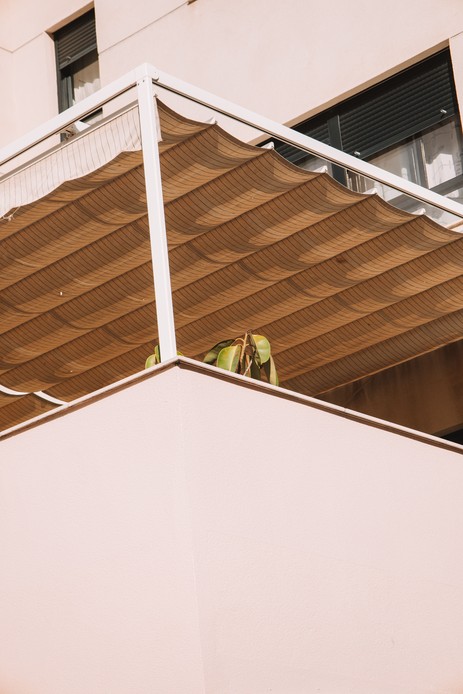 Balcone schermato in alto da una tenda in tessuto a onde nei toni del sabbia, contro una parete chiara
