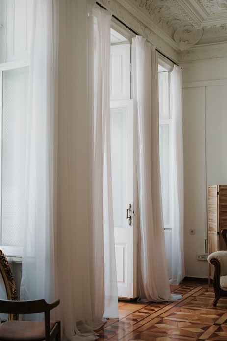 Finestre e porta alte in un interno d'epoca, vestite da tende bianche leggere fino a terra, con pavimento in parquet intarsiato