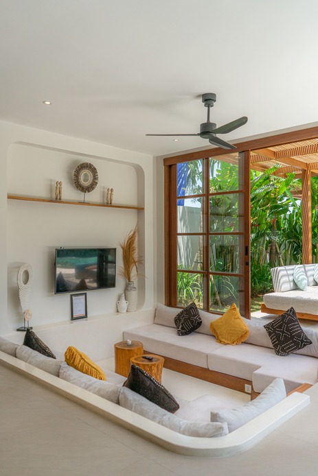 Soggiorno di una villa tropicale con ventilatore a soffitto, ampie porte a vetro aperte sul giardino e divani chiari