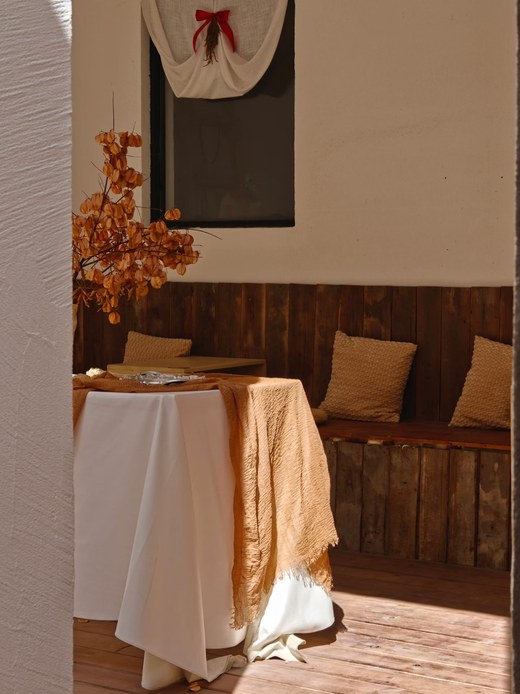 Angolo esterno con tavolo coperto da tovaglia bianca e telo ocra sfrangiato, panca in legno con cuscini color sabbia e rami secchi con bacche arancioni in vaso.