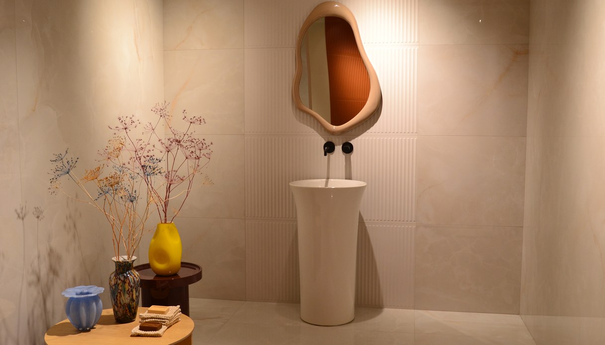 Bagno espositivo con pareti in grandi lastre effetto onice, lavabo a colonna bianco, specchio dalla forma organica e vasi in vetro colorato su tavolini tondi