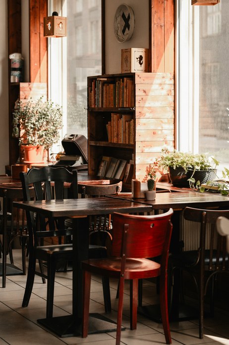 Interno di caffetteria con tavoli scuri e sedie in legno curvato nero e rosso, libreria in cassette di legno, macchina da scrivere d'epoca e piante alla vetrata