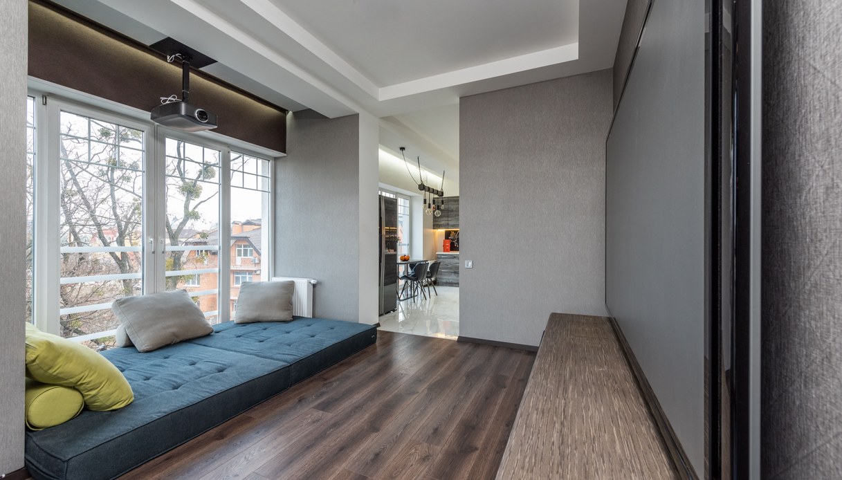 Stanza con proiettore fissato al soffitto, grande telo a parete e materasso imbottito blu con cuscini sotto una vetrata