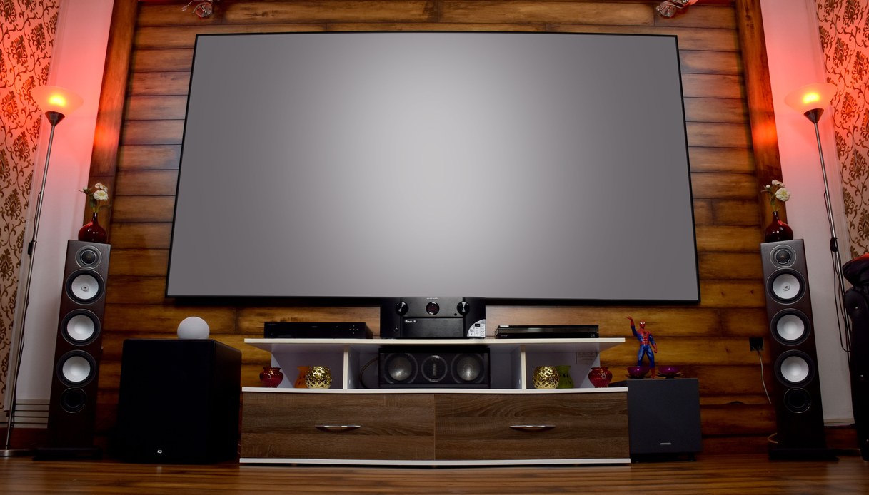 Grande schermo su parete rivestita in doghe di legno, con due diffusori a colonna, subwoofer, amplificatore e lettori su un mobile TV