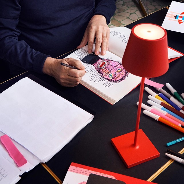 Lampada da tavolo ricaricabile rossa accesa su un piano di lavoro nero, tra pennarelli colorati, fogli e un quaderno di schizzi su cui sta disegnando una persona
