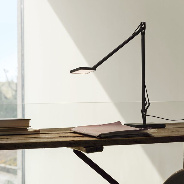 Lampada da scrivania nera a braccio sottile, con testa piatta rettangolare, su un tavolo in legno grezzo accanto a un quaderno e a una pila di libri