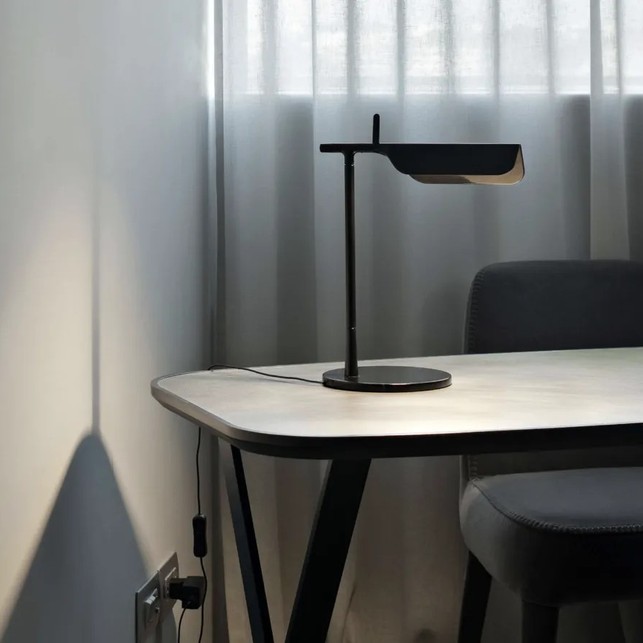 Lampada da tavolo nera con testa a visiera orientabile, appoggiata su un piano bianco davanti a una tenda leggera, con sedia imbottita grigia a lato
