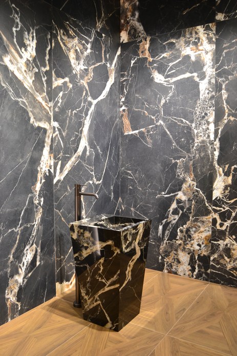 Lavabo monolitico effetto marmo nero con venature bianche e ambra, rubinetteria alta brunita e pareti rivestite con lastre dello stesso materiale su pavimento effetto legno
