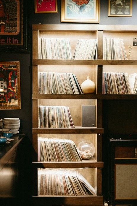 Libreria in legno con vinili conservati in verticale su cinque ripiani, un piccolo diffusore e vasi in ceramica, contro una parete scura con quadri incorniciati