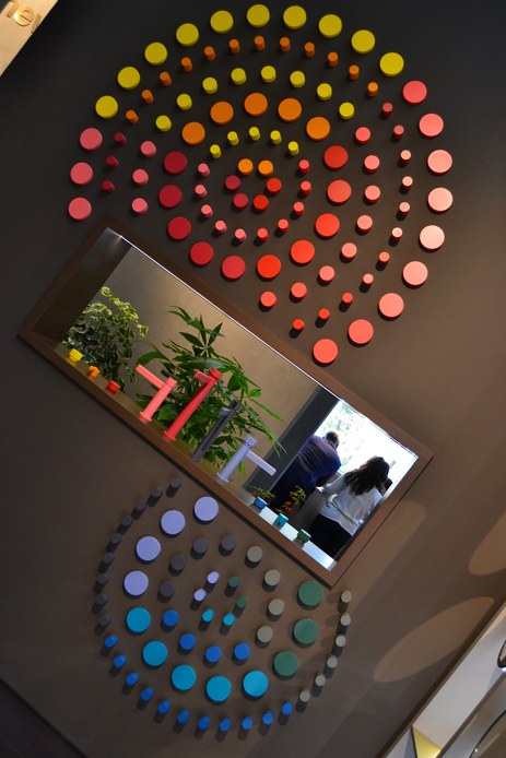 Parete grigia decorata con dischi in ceramica di diverse dimensioni nei toni del giallo, arancio, rosso, azzurro e verde, con uno specchio rettangolare inclinato al centro