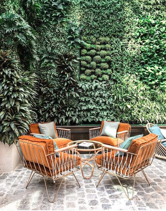 Patio pavimentato in pietra con quattro poltroncine in metallo dorato e cuscini in velluto arancio, tavolino tondo in vetro e una grande parete verde di felci e piante.