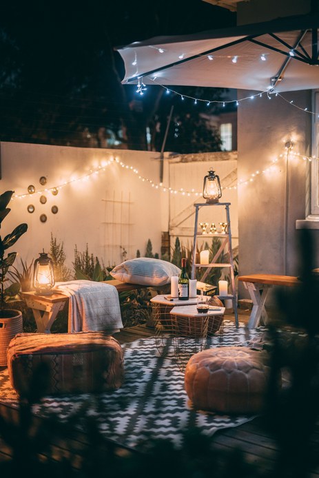 Patio serale illuminato da catene di lampadine e lanterne a batteria, con pouf in pelle, panche in legno, plaid, candele e tappeto a trama geometrica.