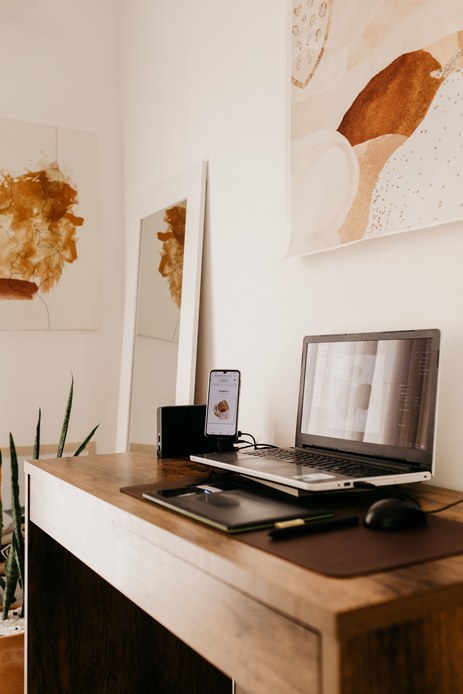 Laptop su rialzo con smartphone in appoggio e tavoletta grafica su un piano in legno, davanti a una parete bianca con stampe astratte dai toni ocra