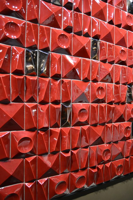 Parete rivestita con piastrelle tridimensionali rosse dalle forme geometriche, tra piramidi, incavi circolari e inserti lucidi scuri