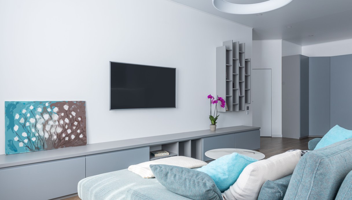 Soggiorno luminoso con TV a parete sopra un mobile basso grigio, divano angolare in tessuto azzurro con cuscini e plaid, tavolino rotondo in marmo
