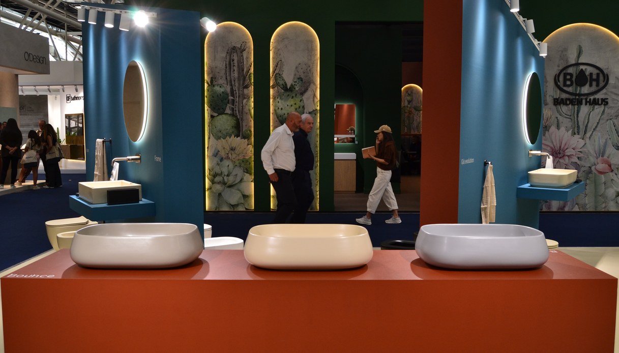 Stand fieristico di arredobagno con tre lavabi da appoggio in ceramica su un banco color terracotta, pareti in verde e blu petrolio e decori botanici retroilluminati